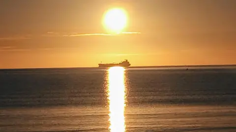 The silhouette of a tanker sails from right to left. It sits perfectly in line with the setting sun and it's reflection on the calm seas. The clear skies are yellow as the sun heads towards the horizon.