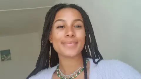 Darwin Alford Darwin Alford smiles at the camera while taking a selfie inside a room painted white. She wears a fluffy pale purple cardigan and a green beaded necklace, her braided hair worn loose over her shoulders. 