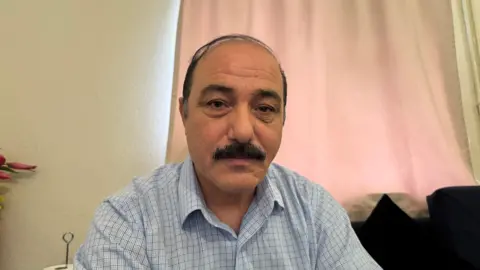 A man in his 50s from Afghanistan looks directly into the camera. He has dark hair in a combover style, and a neatly trimmed dark moustache. He is wearing a light blue checked shirt, and the blue sofa he is sitting on can be seen in the background. 