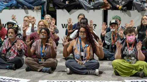 Climate activists sitting on the floor wearing tape over their mouths, with "Pay Up" written on the tape. 