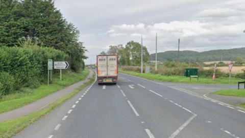 A view of the A46 towards Evesham with a lorry driving on the left and fields and trees in the distance on the right