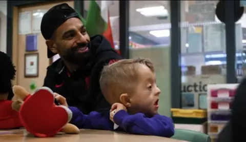 Young Isaac clutches a plush LFC toy while at his class table with Mohamed Salah sitting beside him