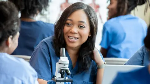 A young healthcare worker is leaning on a desk with a microscope on it, and smiling and chatting to the person opposite her. She has long, dark brown hair, wears blue overalls and is surrounded by others in the same clothing.