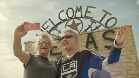 Harper Steele, wearing black glasses with her hair swept up, takes a selfie with Will Ferrell, who has short grey hair and is wearing black sunglasses