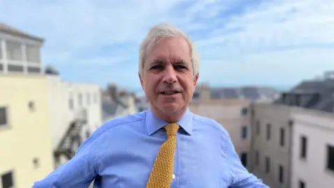 Christopher Beaumont pictured wearing a light blue shirt with a yellow tie and stood over a balcony.