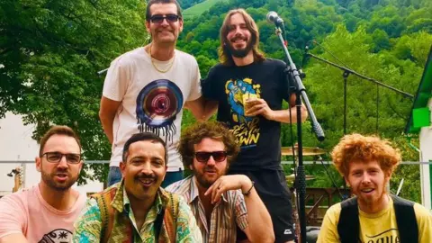 Four band members, all male, are sitting on the edge of an outdoor stage as two other male band members stand behind them. They are stood in front of microphones with a drum kit. In the background there are lots of trees with deep green leaves. 