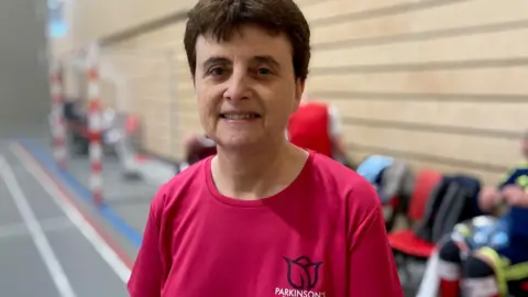 Julie Norris is a woman in her sixties wearing a pink top. She has dark hair and is smiling 