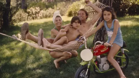 Alamy Sir Roger Moore lies on a hammock. He is wearing swimming trunks and has his arm outstretched above him. He is smiling and looking relaxed. His wife Luisa is crouching down alongside the hammock. She is also wearing swimwear and has a towel wrapped around her hair. She looks content. Sir Roger's young son Geoffrey is sitting on his lap. He has dark brown hair and is looking at the camera apprehensively. Sir Roger's daughter Deborah is sitting on a small green motorcycle beside the hammock. She also has dark brown hair and is wearing a pale blue dress. She has a half smile on her face.