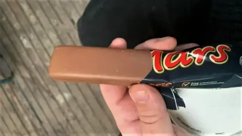 A Mars bar, with no ripple, being held in someone's hand. The wrapper is partially off.