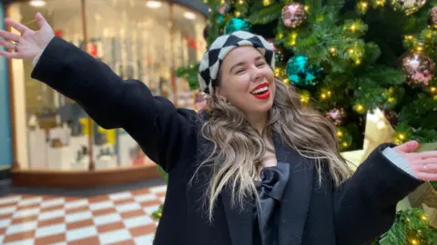 Amy Langston is wearing a black coat and a top with a black ribbon. She has  long brown hair and is wearing a black and white checkered beret on her head. Amy is standing in front of a Christmas tree and is smiling.