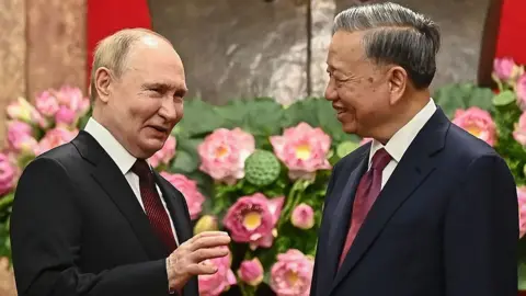 Vladimir Putin gestures while smiling as he interacts with a smiling To Lam, both wearing suit and tie against a backdrop of pink and green flowers, in Hanoi on 20 June
