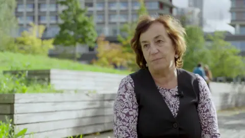 Professor Penny Bernstock in a purple and white floral blouse and dark waistcoat. She is sat on a bench in the Queen Elizabeth Olympic Park.