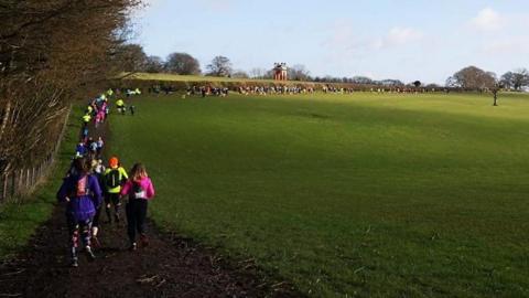 A long line of runners snaking around the edge of a grassy field and disappearing into the distance