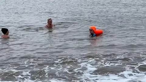 Andrew Turner/BBC Pam Spychal, with an orange hat and float, swimming in the sea with two of her friends, to raise money for charity. Pam is on the right of the image.