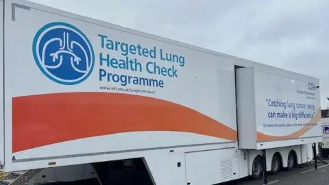 A large white and orange lorry trailer with the words 'Targeted Lung Health Check Programme' painted in blue on the side, with an illustration of a pair of lungs next to it, parked up in a Tesco's car park in Greater Manchester.