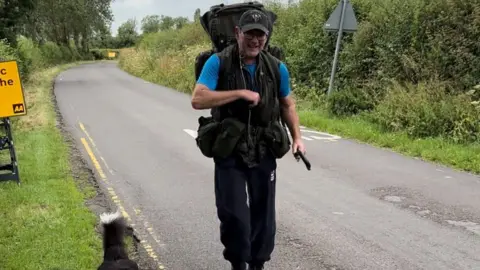 Philip wearing his old army gear and a blu t-shirt and dark cap walking on a road with his dogs