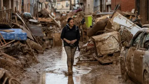 A sombre-looking middle-aged woman covered in mud walks down a flooded street in Valencia which is filled with damaged furniture, kitchen appliances and other household goods after heavy rain and flooding hit large parts of Spain on 2 November, 2024. To the right of the image is A badly-damaged car with no windows.