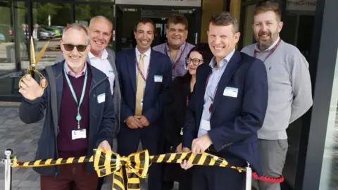 A group of people in front of a ceremonial ribbon, with the man on the left holding some golden scissors