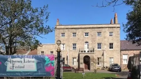 A three-story house made of light brown brick. There are two cars parked in front of the house. There is a banner on the gate to the house which reads 'Alderney Literary Festival'.