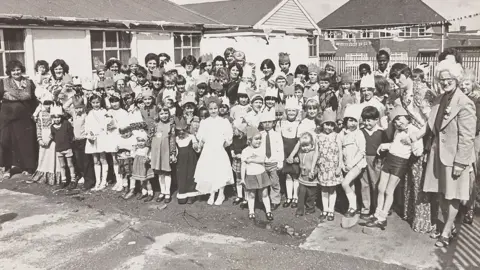 Express & Star/Wolverhampton City Archives A black and white picture of a large group of children of more than 40 children with at least 10 adults among them, The children are party outfits, including dresses and skirts, while one of the boys wears a tie. Some of the children are wearing crown hats. 