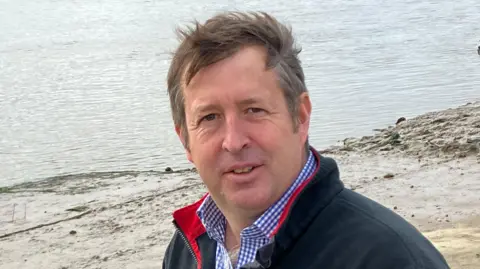 Commodore Henry Walker stands on the old concrete slipway wearing a blue chequered shirt and a dark fleece top, with the water of the River Blyth in the background