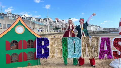 Amanda White/BBC Two women dressed as elves on stilts stand next to a tinsel 'Bridmas' sign