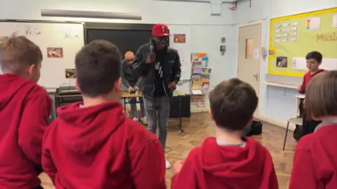 Rapper Smack Jay stands in the middle of a classroom with a microphone in his hand. A number of schoolchildren wearing red school hoodies are watching him.