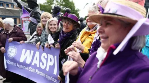 Women in bright purple clothing stand in a line behind a Women Against State Pension Injustice campaign sign