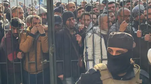 An armed and masked man stands inside a gate, while a crowd of other men press against the gate.