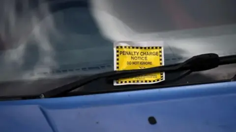 A close-up of the windscreen of a blue vehicle with a sign reading: "Penalty charge notice - do not ignore" stuck under the windscreen wiper