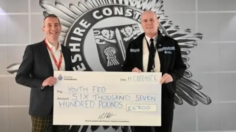 Two men - one in a suit jacket and the other in a police uniform - hold a large novelty cheque which reads 'Youth Fed, £6,700'. 