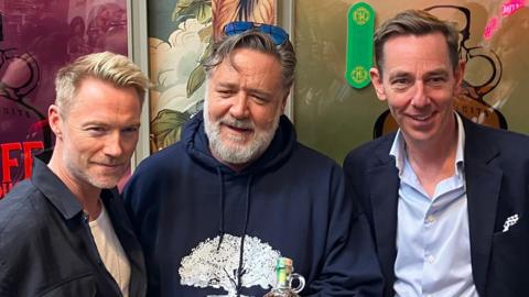 Russell Crowe, Ronan Keating and Ryan Tubridy pictured together in front of an artful wall with different coloured designs at the Muff Liquor Company. Keating is one the left, wearing a dark shirt and white t-shirt with short, cropped blonde hair. Crowe is in the middle with a blue hoodie and sunglasses perched on top of his head. Tubridy is on the right wearing a navy suit and light blue shirt. 