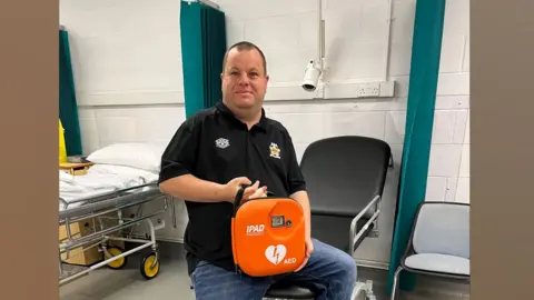 Cambridge University Hospitals NHS Foundation Trust Paul Harrison wearing a black polo-shirt and perched on a black hospital trolley, holding an orange box containing an automated external defibrillator. He has short dark hair and is looking at the camera. 