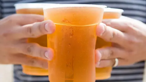 Close-up image of three pints of beer in plastic cups being carried by a man. The man's hands are visible either side of the pints, holding them together. He is wearing a stripy t-shirt