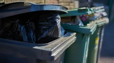 A black wheelie bin is in the foreground with the lid slightly open, exposing a black bin bag. Next to it, in the background, are two green recycling bins, also with the lids slightly open. This is exposing rubbish.