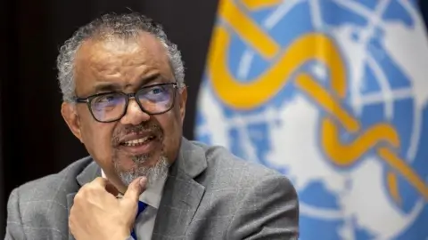 World Health Organization director-general Tedros Adhanom Ghebreyesus shot from chest height up, holding his left hand to his chin, with a UN flag in background