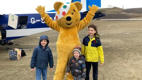 Pudsey the bear with a colourful polkadot bandage over one eye standing with three children in front of a small blue and white plane on the Fair Isle landing strip.