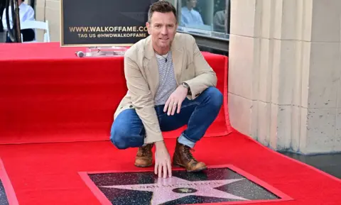 Getty Images British actor Ewan McGregor touches his star during his Hollywood Walk of Fame Star ceremony in Hollywood, California. He is wearing brown boots, jeans and a beige jacket.