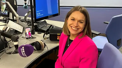 BBC/Julia Moore Young woman with brown hair and a bright pink jacket smiling and looking at the camera in a radio studio and in front of a purple microphone with BBC Radio Surrey logo on it. 