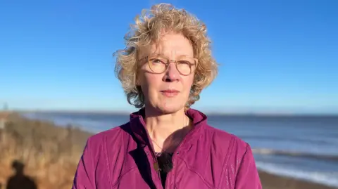 Deborah Hawksley stands on the edge of the cliff at Skipsea. The sea is visible in the background with blue sky and grassed landscape. Ms Hawksley is wearing a dark pink jacket and gold necklace. She has light coloured glasses and blonde curly hair.