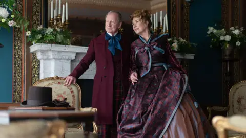 Mark Mainz Actor Bill Paterson is dressed in a period costume consisting of a long burgundy jacket, blue cravat and tartan trousers. He is leaning against an old-fashioned chair while standing in front of a white marble fireplace and mirror. Next to him is Julie Cadzow, also dressed in a costume. Her outfit is a burgundy and blue tartan dress and jacket.