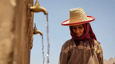 A person looks down while wearing a sunhat with taps pouring water into containers (out of frame) with a blue sky in the background, in Morocco in June 2024