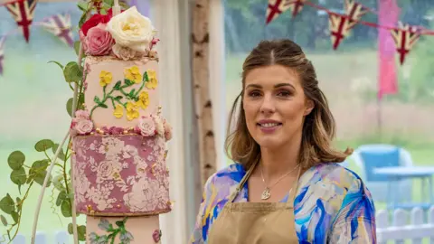 Love Productions Georgie Grasso stood in the Bake Off tent. She is wearing a beige apron and underneath it has a blue, pink and white marbled top. She is looking at the camera with her brown and blonde hair half tied up and half down. There is a cake to the left of her, which is a three tiered cake, in different shades of pink with flowers adorned on it. 