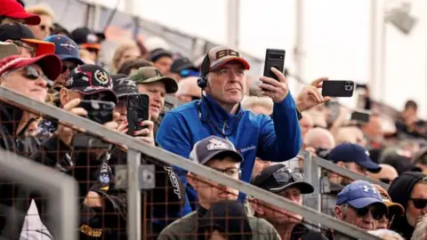 Fans wearing TT caps sit in the grandstand. Many are filming the action on their phones.