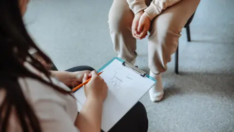 Getty Images A child sits on a chair with a drawing on a clipboard. A second person's legs are visible. They are sitting on a chair facing the child. 