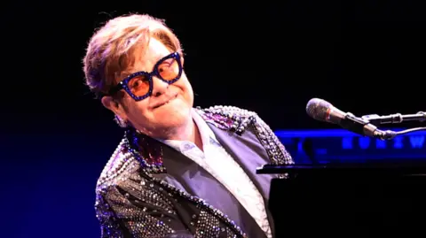 Elton John plays piano while dressed in a purple sequinned outfit