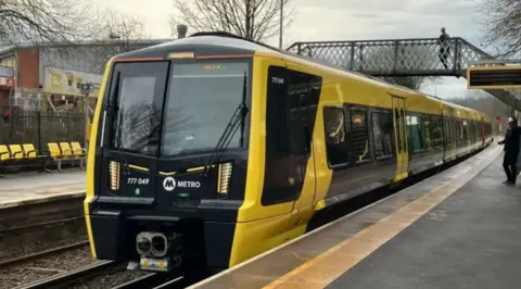 A Merseyrail black and yellow electric train on the track. A bridge over the platform can be seen above the train with a man walking across the other side. One man can be seen standing on the train platform at the bottom looking towards the train.