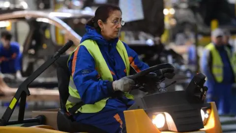 A woman driving a vehicle inside  a factory, wearing a hi-vis vest on top of work overalls. 