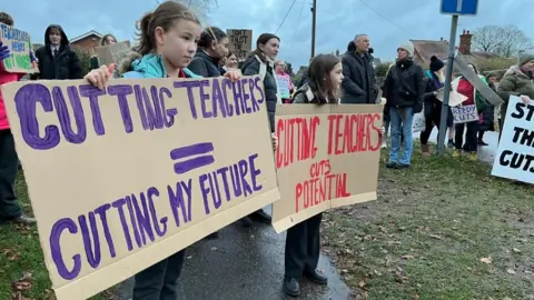 Shaun Whitmore/BBC Children and parents with banners and placards outside Reepham High School and College. Placards read: "Cutting teachers = cutting my future" and: "Cutting teachers cuts potential".