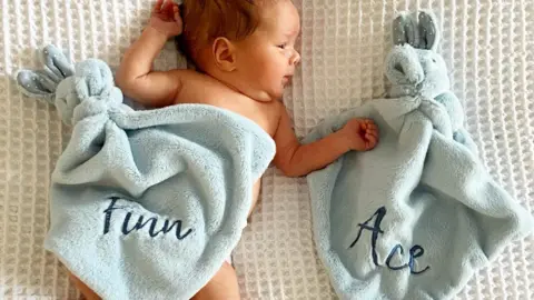 Baby Finn lying under a blanket with his name on, next to a blanket that has his brother's name on